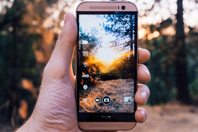 A hand holding a smartphone in a wooded landscape, with sunlight and trees shown more clearly within the phone screen.